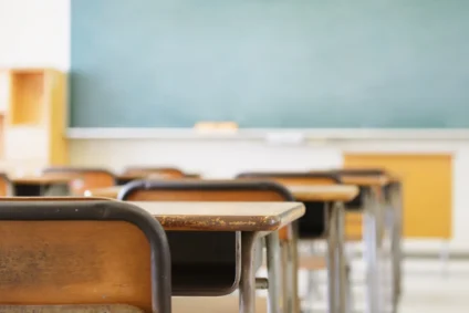 Banchi scolastici vuoti in legno e metallo in un'aula con lavagna verde sullo sfondo sfocato.