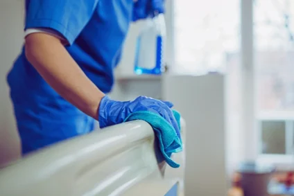 Operatore sanitario con guanti blu esegue la disinfezione delle superfici di un letto d'ospedale con un panno.