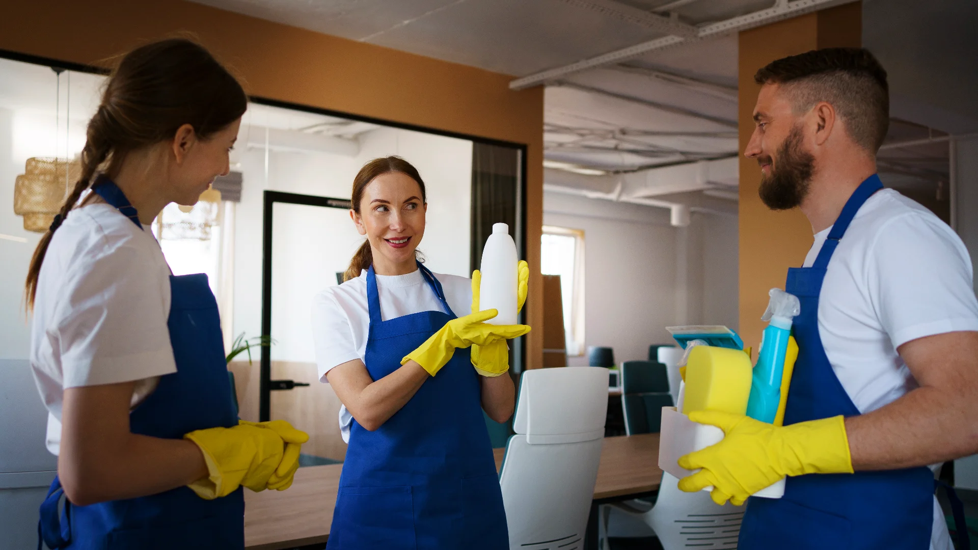 Addetti di un'impresa di pulizie professionali con grembiule blu e guanti gialli confrontano prodotti detergenti in ufficio.