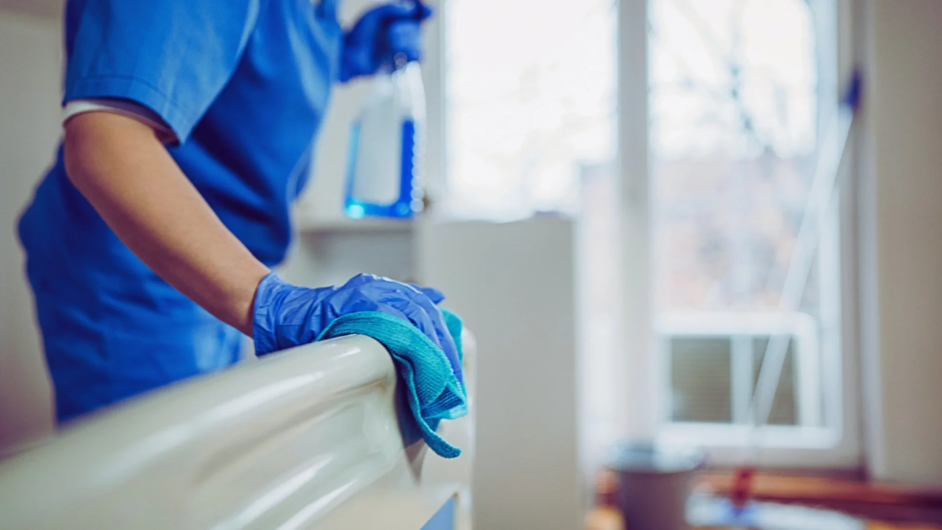 Operatore sanitario con guanti blu esegue la disinfezione delle superfici di un letto d'ospedale con un panno.